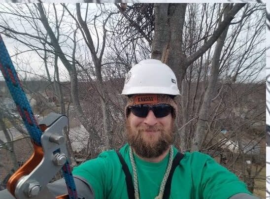 Ronnie in a white climbing helmet and green FLATS shirt, roped in up a tree